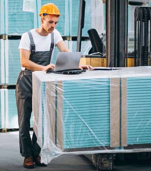 Warehouse worker with safety helmet using laptop in industrial setting.
