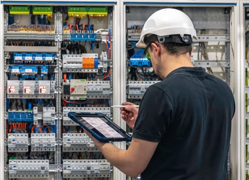 Electrical technician using tablet to inspect electrical control panel.