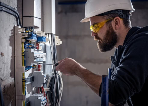 Electrician working on electrical panel with tools and safety gear.