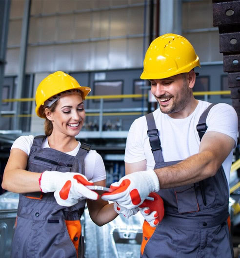 Two factory workers in yellow helmets sharing a tablet in a manufacturing plant.